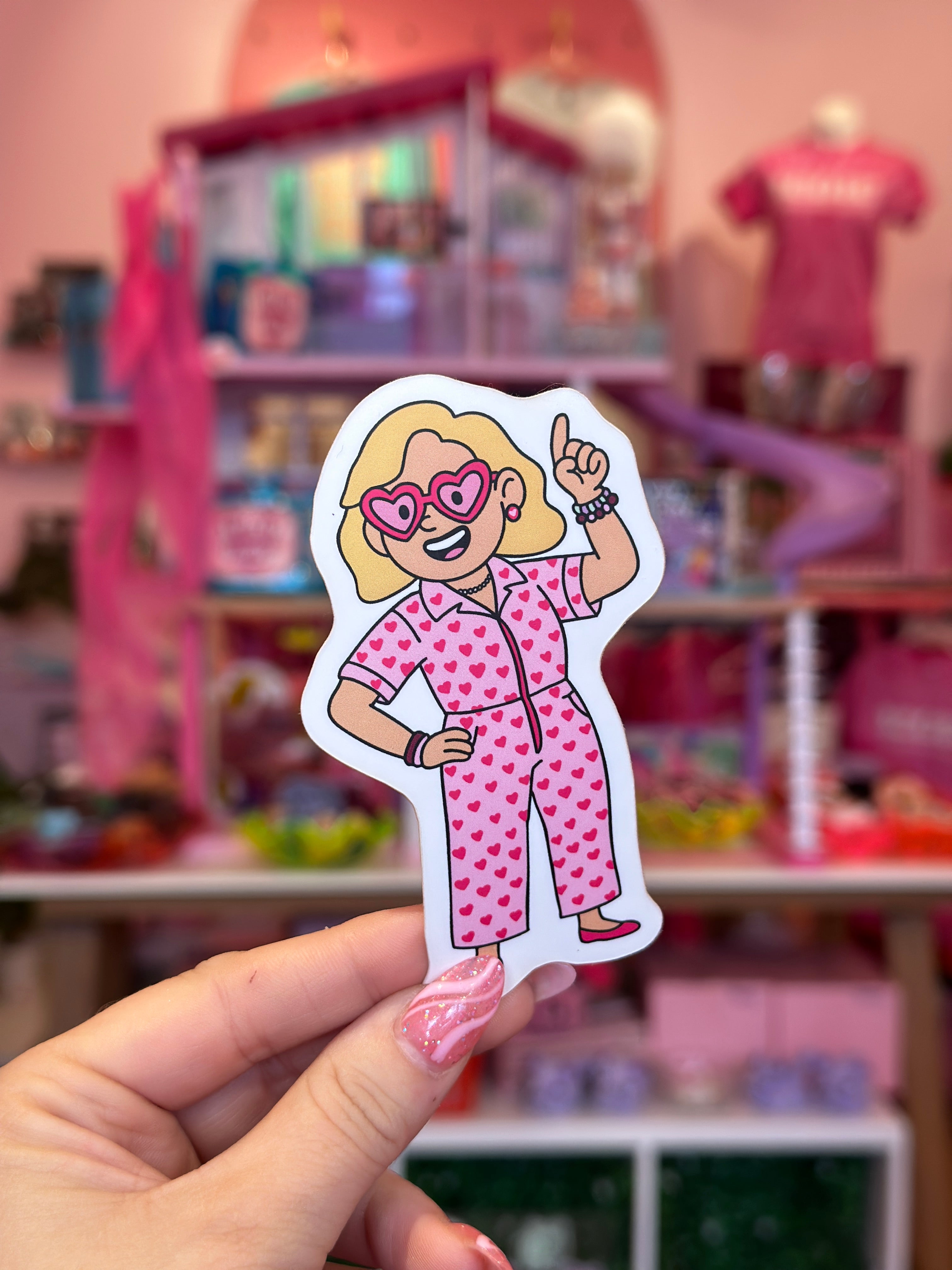 Hand holding a sticker of a character in pink pajamas against a blurred toy store background.