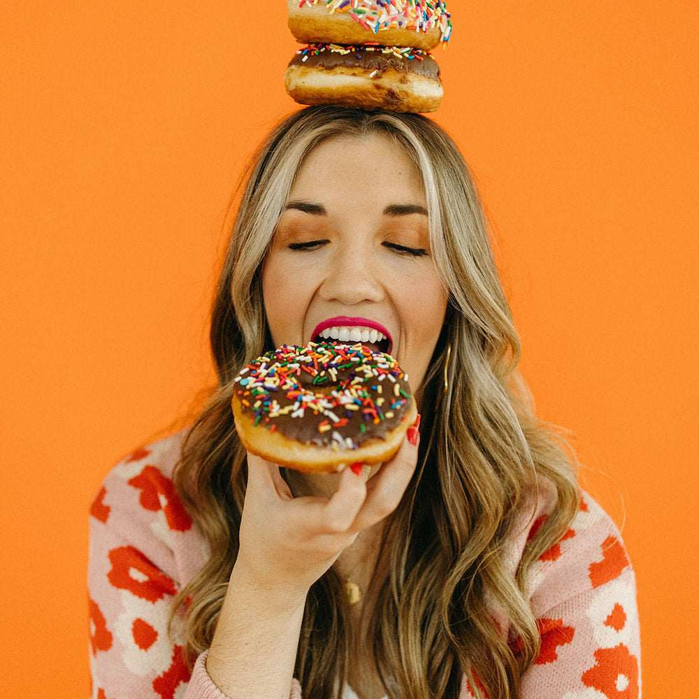 Close up photo of woman biting into a chocolate covered donut while balancing two donuts on her head. She has brown hair and is wearing red lipstick and a floral sweater. She she sitting in front of a bright orange backdrop. 