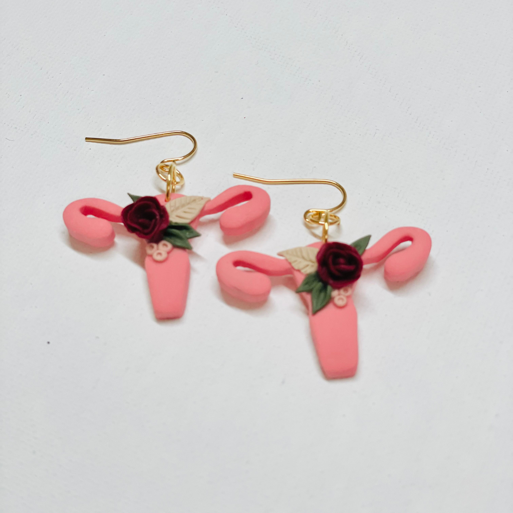 A pair of earrings are shown lying against an off-white background. The earrings are pink polymer clay earrings in the shape of a uterus, with polymer clay floral arrangements placed just off center on top. 