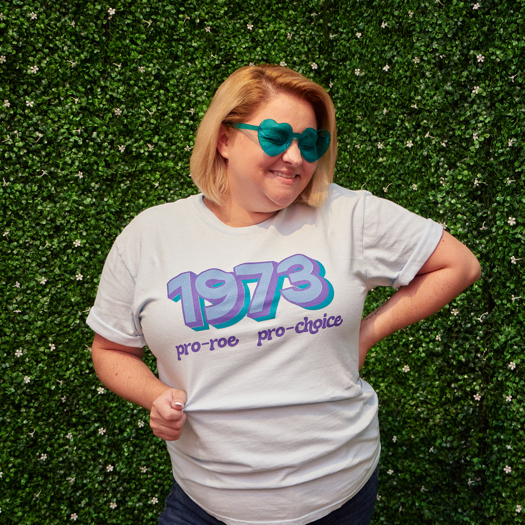 Woman wearing light blue t-shirt that says "1973 Pro Roe, Pro Choice" in light blue, purple, and teal retro-style fonts. Shirt was designed by Woman-Owned Wallet, a feminist gift shop in Louisville KY.