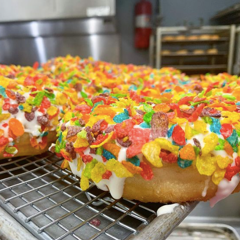 Two donuts covered in white icing and colorful Fruity Pebbles cereal sitting on a cooling rack. Donuts made by Hi-Five Doughnuts, one of the featured businesses on the Woman-Owned Walking Tour of Nulu and the Woman-Owned Directory.