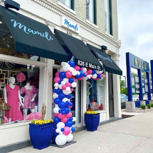 Photo of the Mamili storefront featuring a white painted brick building with black awnings, styled mannequins in the windows, and a pink, white, and blue balloon arch over the front door. Mamili is one of the shops featured in the Woman-Owned Directory.