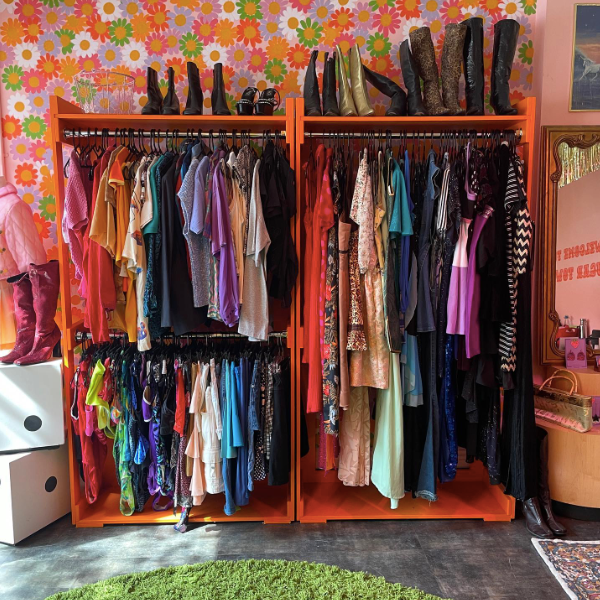 Bright orange clothing racks with a variety of colorful vintage clothing and boots. The wall in the background has a whimsical floral printed wallpaper. This is Sugar Town Vintage, one of the businesses featured in the Woman-Owned Directory.