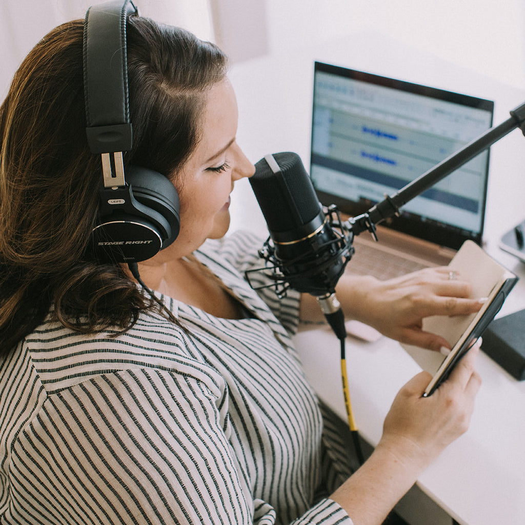 Amanda Dare, owner of Woman-Owned Wallet, is sitting at a desk wearing headphones and speaking into a microphone while looking at the script for Woman-Owned Wallet: The Podcast. WOW is a feminist gift shop in Louisville KY.