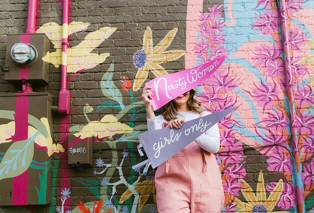 Woman standing in front of a colorful, floral mural in Louisville Kentucky holding up two pennants in front of her face. These pennants are from Woman-Owned Wallet, including one that says "nasty woman" and other other and "girls only."