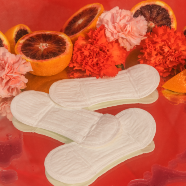 Three period liners laying in front of pink and red carnations and blood oranges on a red background. The liners are from a period care brand named August.