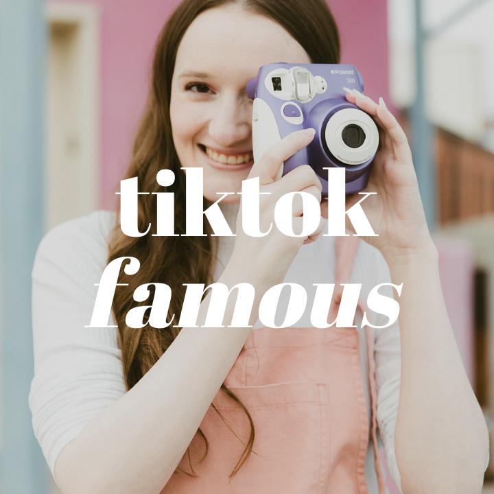 Woman smiling at the camera while holding a purple Polaroid camera up to her left eye. She is wearing a set of pink overalls and a long sleeve t-shirt. The words "TikTok Famous" are typed in large white text in the middle of the photo.