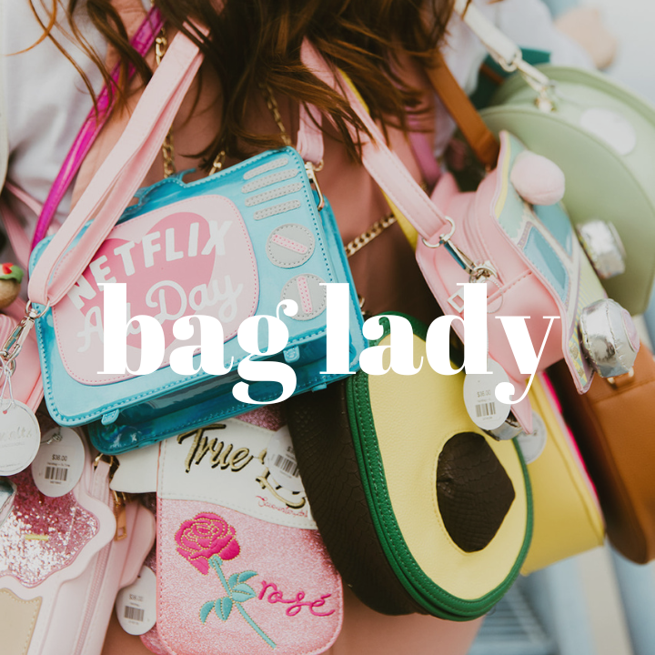 Close up of woman wearing multiple novelty purses across her body, including bags that look like a television set, a champagne bottle, an avocado, a taco truck, etc. The words "Bag Lady" are typed in large white text in the middle of the photo.