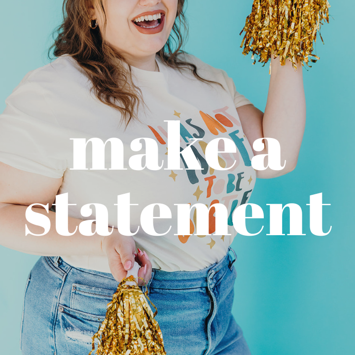 Woman smiling and holding gold pom-poms in her hands while wearing a bold graphic tee and blue jeans. The words "Make A Statement" are displayed in large white text in the center of the photo.