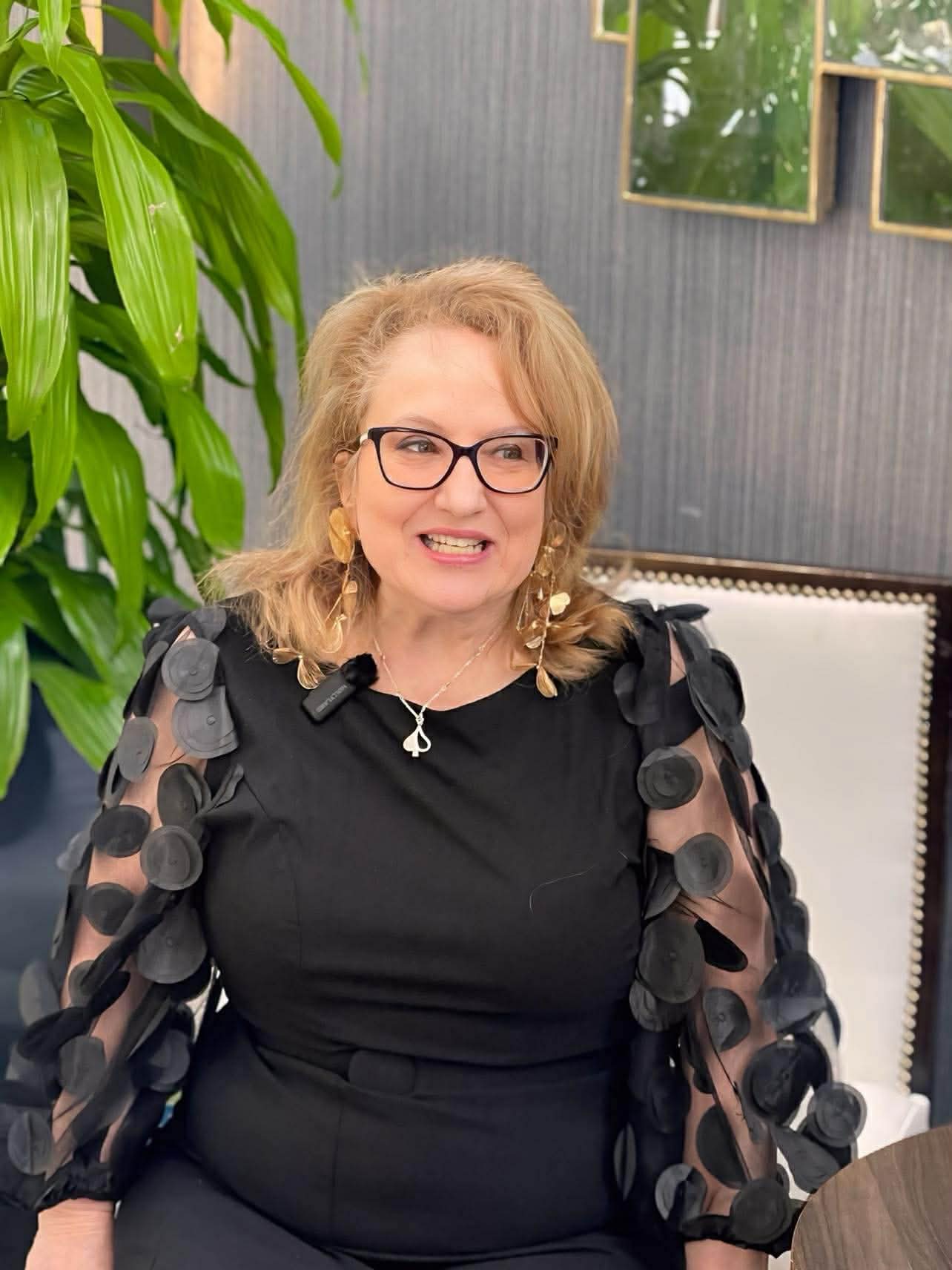 Woman wearing glasses and a black outfit with floral details, sitting in an indoor setting.