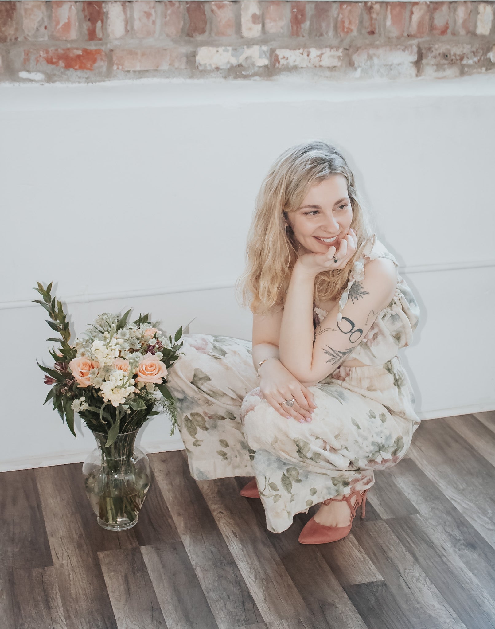 Woman in a floral dress sitting on a wooden floor with a vase of flowers next to her.