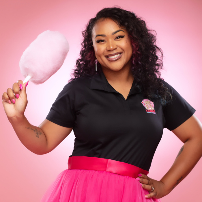 Business owner Brandi wearing black polo with logo and hot pink skirt holds cotton candy stick with right hand and has left hand on hip. Brandi is smiling.