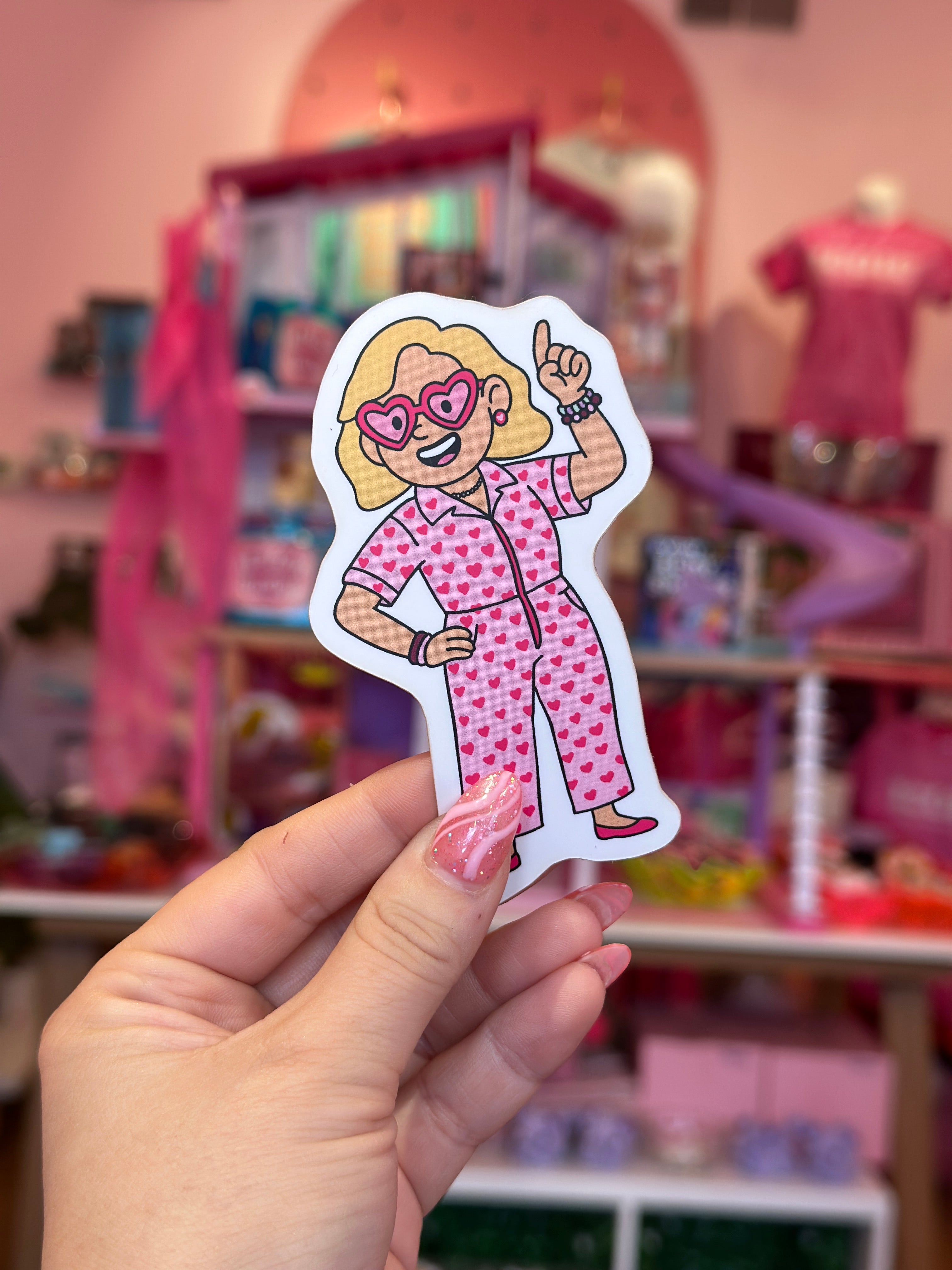 Hand holding a sticker of a character in a pink outfit with a blurred toy store background