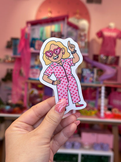 Hand holding a sticker of a character in a pink outfit with a blurred toy store background