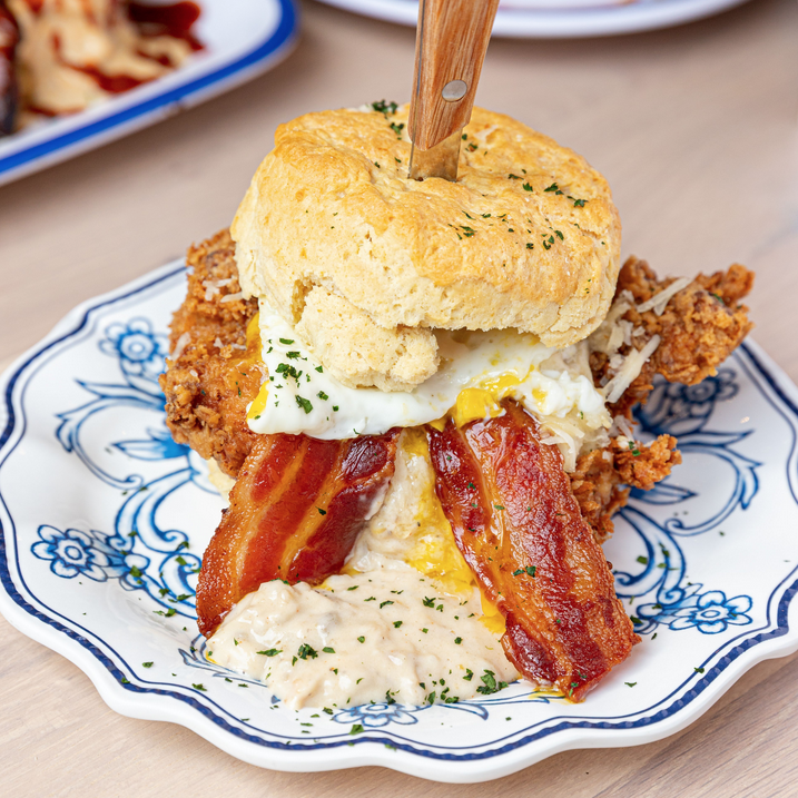 Loaded breakfast biscuit sandwich featuring fried chicken, a fried egg, crispy bacon, and gravy on a white and blue china plate. The breakfast sandwich is from Biscuit Belly, part of the Woman-Owned Wallet tour of Nulu and the woman-owned directory.