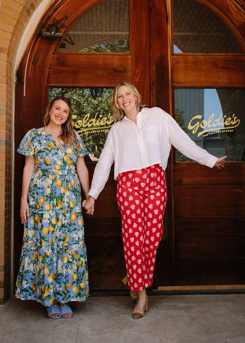 Two women standing in front of a wooden door with 'Goldie's' sign, one in a floral dress and the other in a white shirt and red polka dot pants.