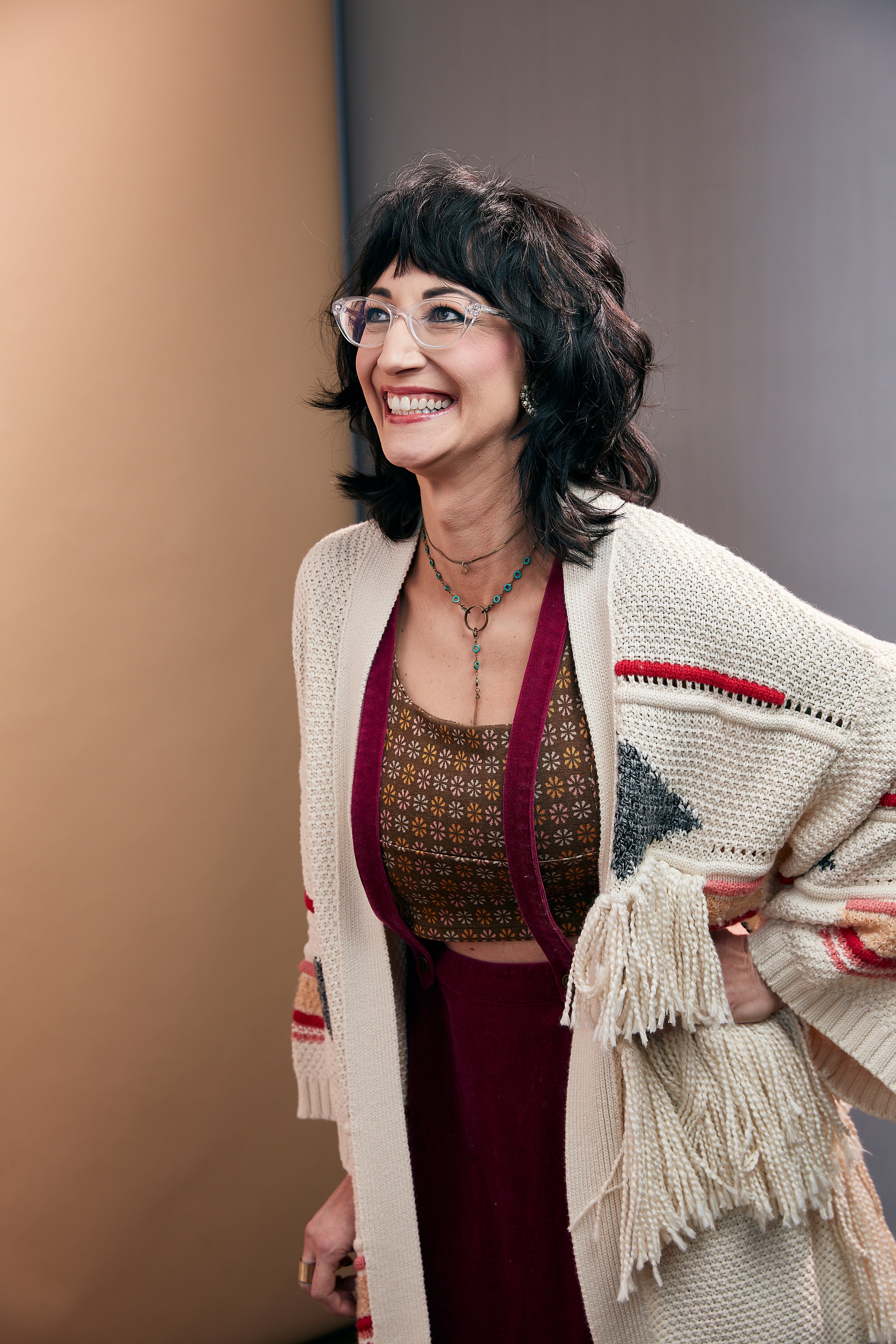 A smiling woman with dark hair and glasses is wearing a cream knit cardigan with fringe and red geometric accents over a patterned brown top and burgundy dress, posed against a neutral studio background.