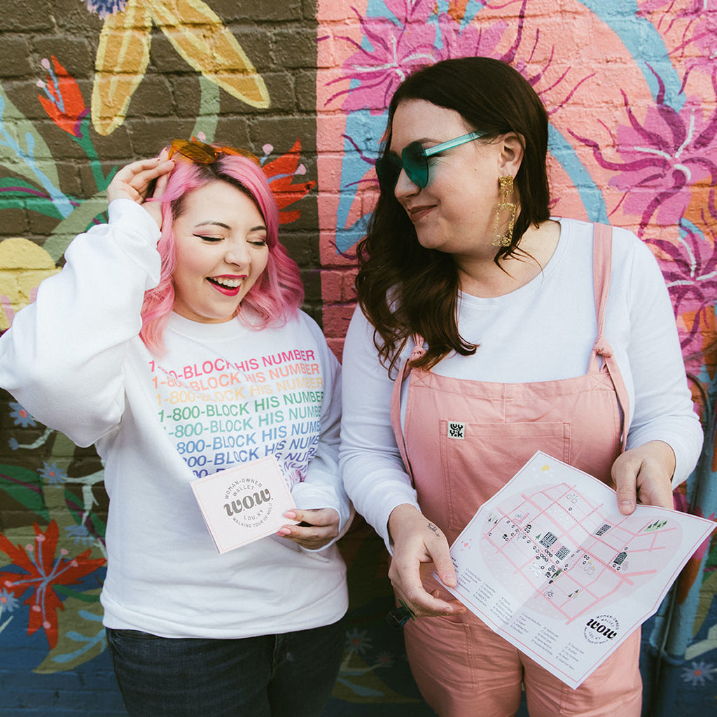 Woman with pink hair wearing a white sweatshirt standing next to a woman with brown hair wearing a white long sleeve shirt with pink overalls. The women are both looking at a map of the Nulu Woman-Owned Walking tour hosted by Woman-Owned Wallet.