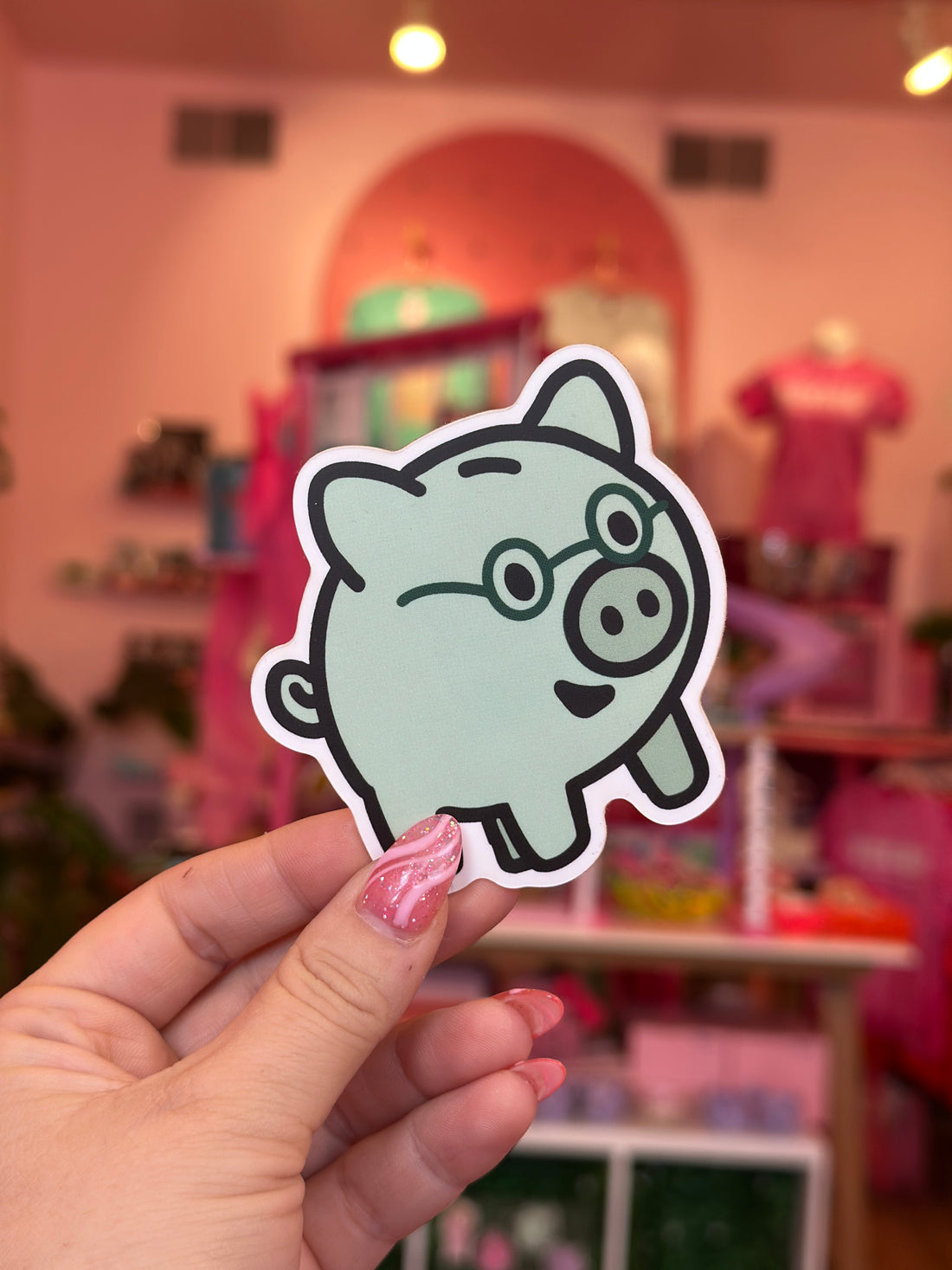 Hand holding a cartoon sticker of a mint green creature with a blurred indoor background