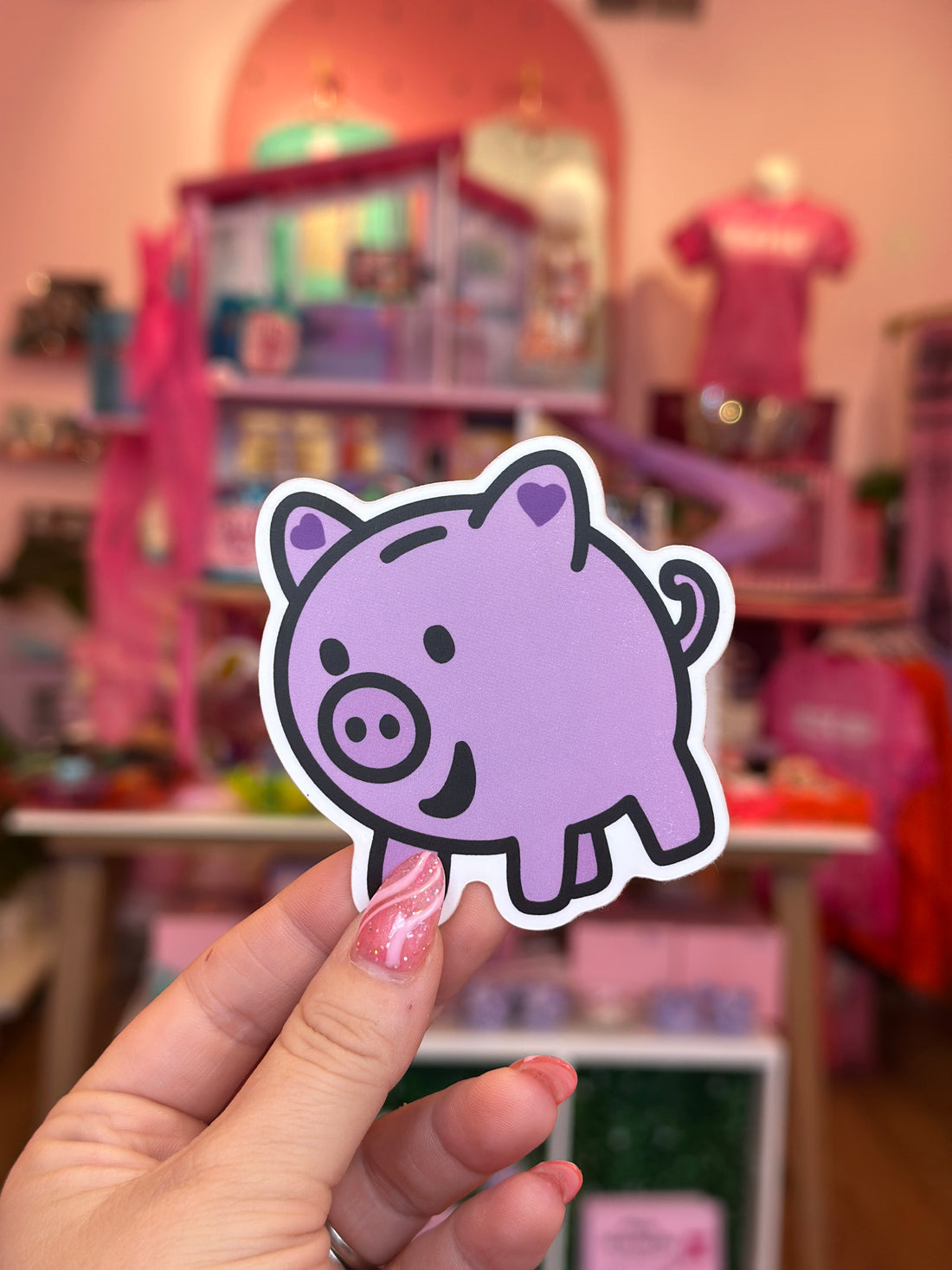 Hand holding a purple cartoon character sticker in front of a blurred toy store background