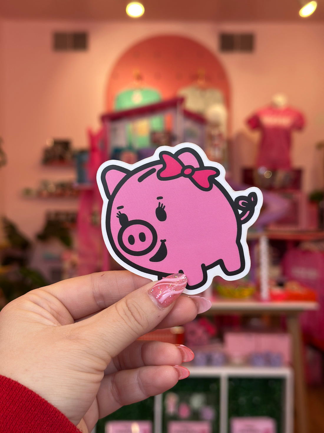 Hand holding a pink cartoon pig sticker with a blurred indoor background