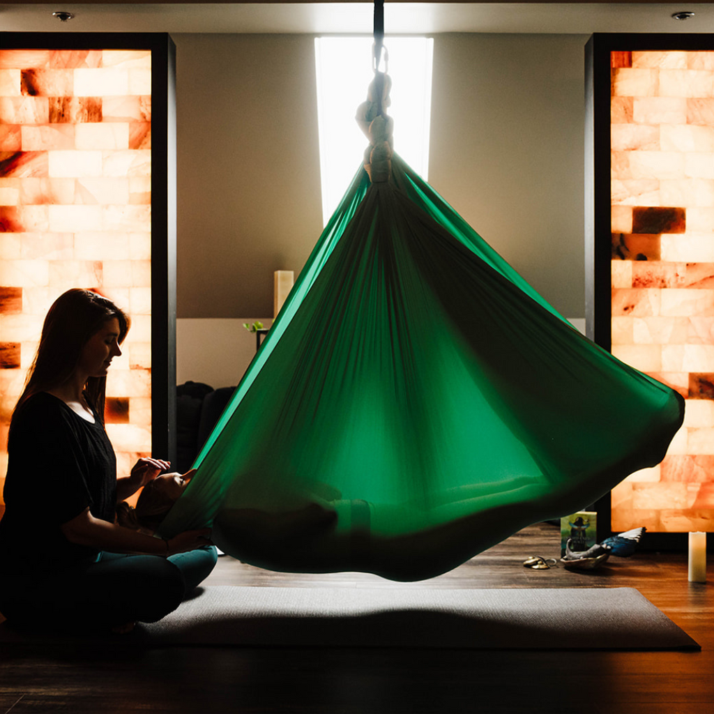 In a low lit room, a woman sits on the floor with her hand over the forehead of a person lying in a green hanging hammock. There is warm natural light coming from the window behind the green hammock. 