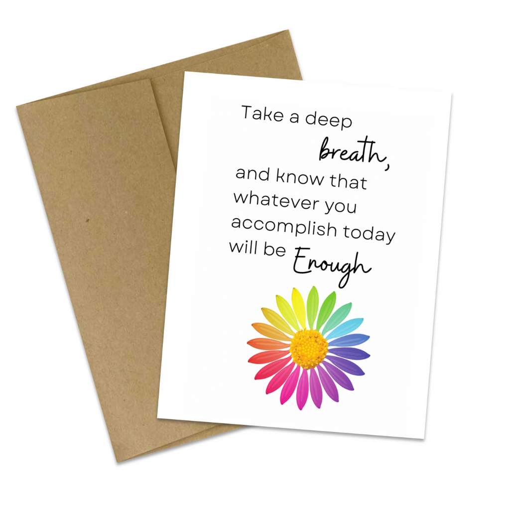 Product photo shows a white greeting card with the words "Take a deep breath, and know that whatever you accomplish today will be enough" featured in the top half of the card, and a daisy with rainbow petals slightly off-centered on the bottom half of the card. Behind the card at a slight angle is a brown paper envelope. 