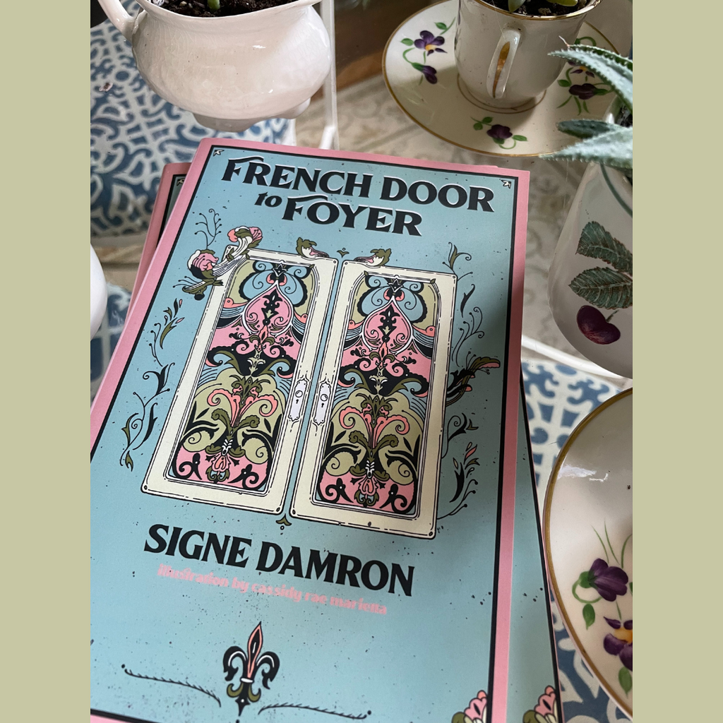 A book rests on a clear table surrounded by dishes. The book has a blue cover with pink trim. The title is boldly reading at the top, "French Door to Foyer" and features a colorful set of French doors adorned with floral paterns. The author's name  "Signe Damron" boldly sits underneath the doors in matching font as the title. a fleur de lis design rests at the bottom of the front cover under the author's name. 