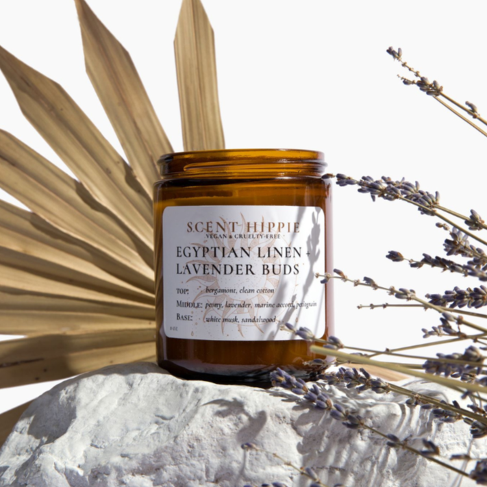An amber glass jar labeled "Scent Hippie Egyptian Linen - Lavender Buds" sit on top of a white rock behind a sprig of lavender with a dried frond in the background, adding dimension to the plan white backdrop.  