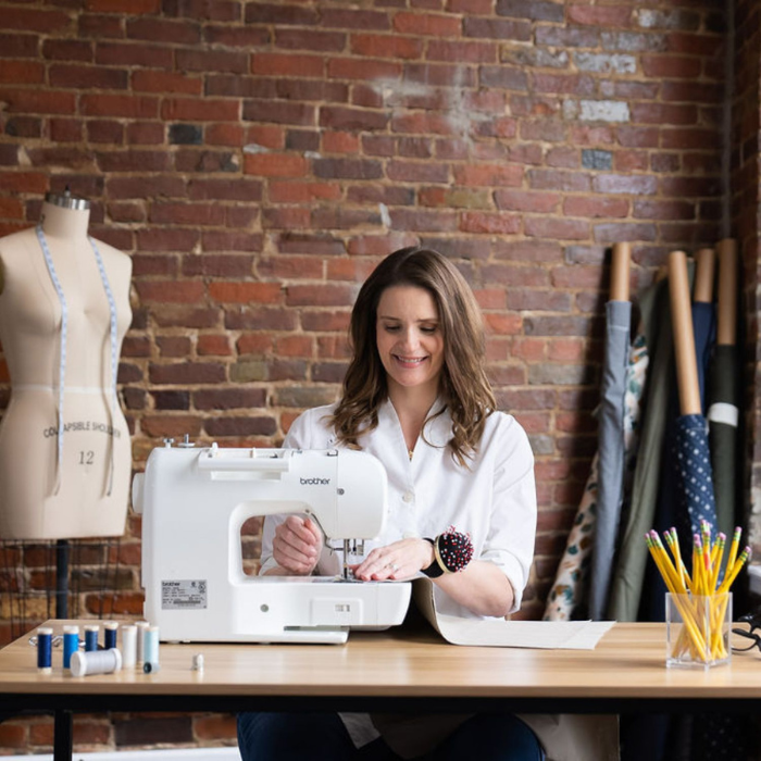 Lyndsey Stanfill sitting at a wooden table cheerfully sewing pieces of fabric together. In the background there is a mannequin and bolts of fabric in front of a brick wall. Lyndsey owns Made Stitch Co. which is featured in WOW's Woman-Owned Directory.