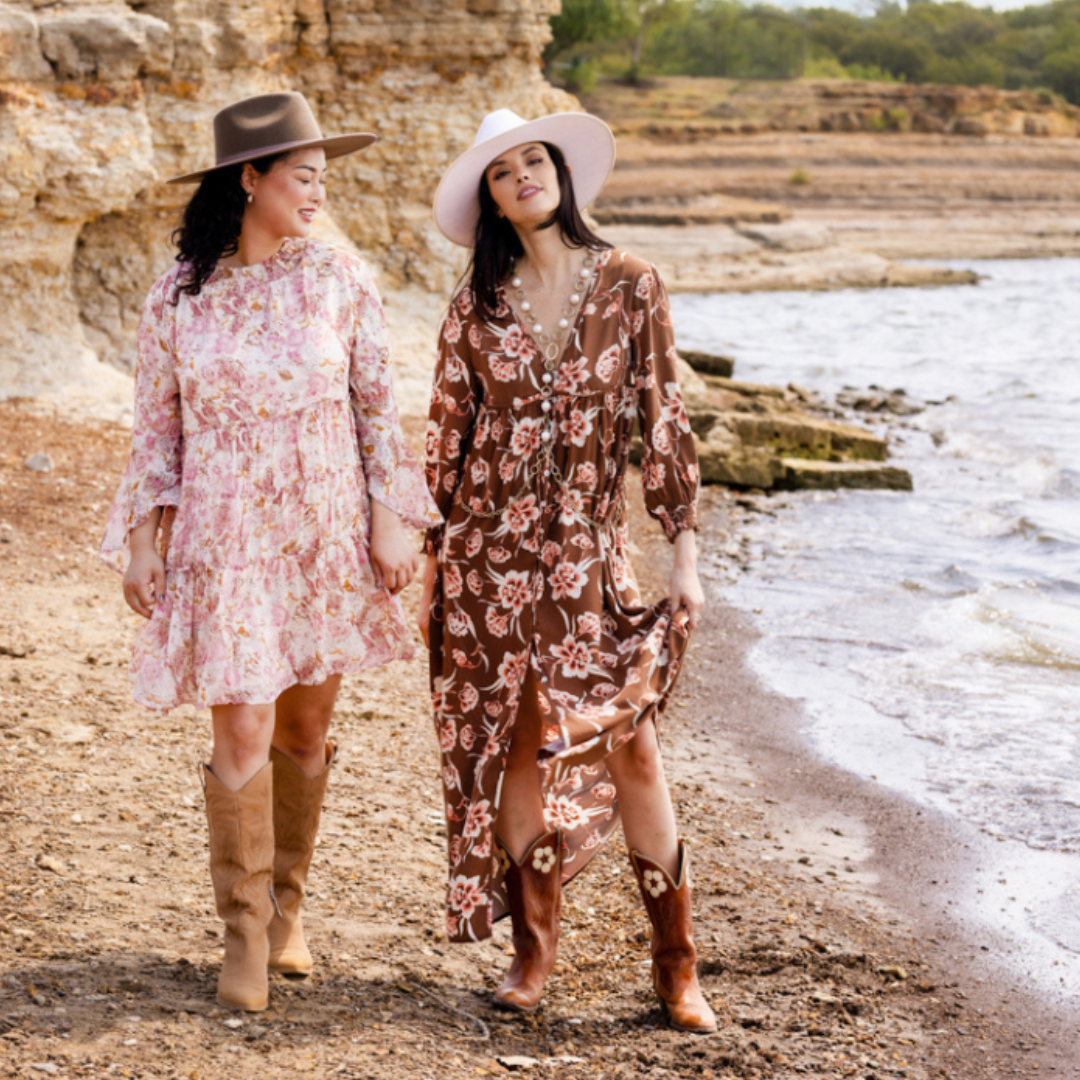 Two women are pictured standing on a rocky shoreline wearing hats, floral dresses, vintage jewelry, and cowgirl boots