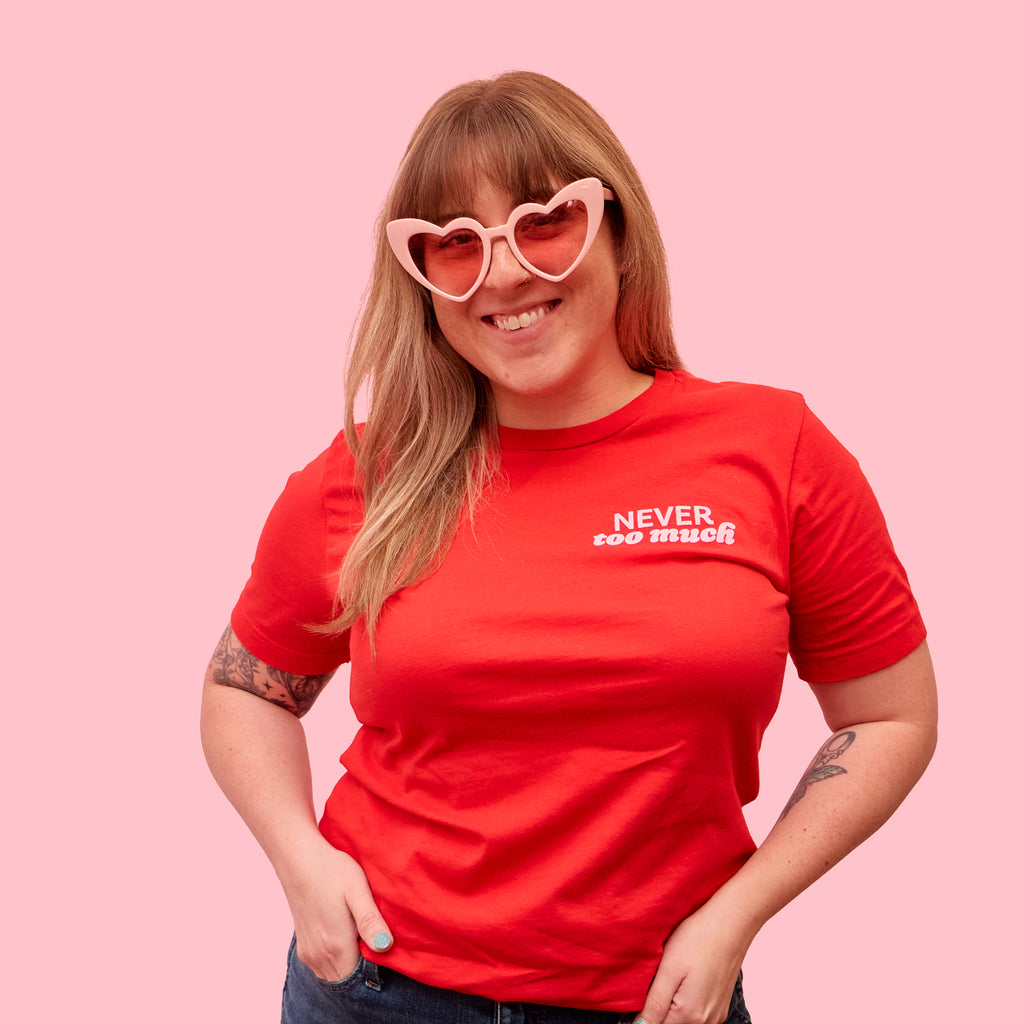 Woman wearing a red t-shirt that says "never too much" in light pink text on the left pocket area. This cute graphic t-shirt was designed by Woman-Owned Wallet, a feminist gift shop in Louisville KY.