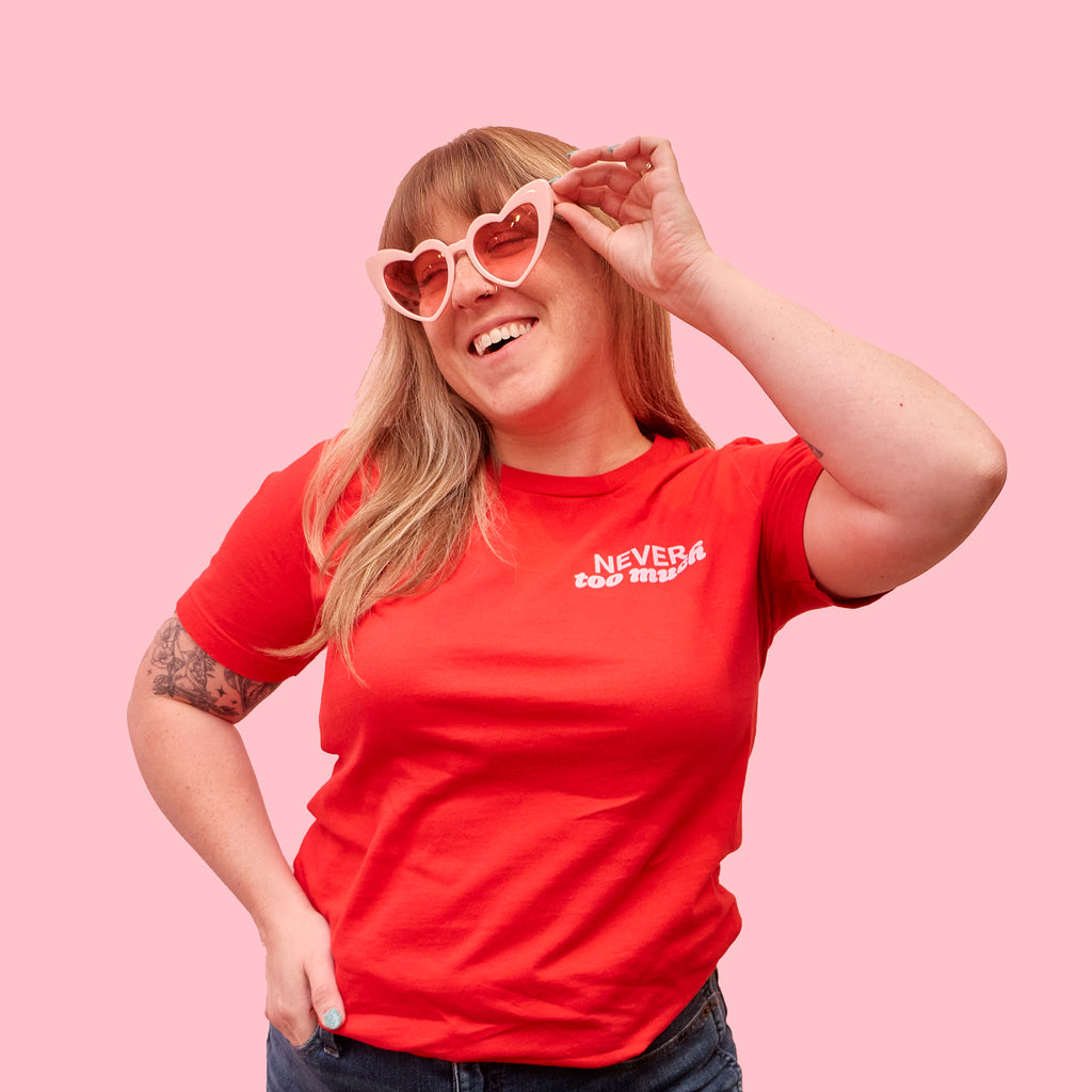 Woman wearing a red t-shirt that says "never too much" in light pink text on the left pocket area. This cute graphic t-shirt was designed by Woman-Owned Wallet, a feminist gift shop in Louisville KY.