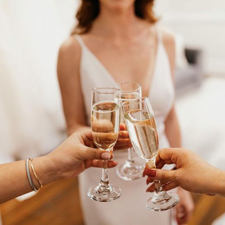 Woman wearing a beautiful white wedding dress and doing a champagne "cheers" with two other people. This photo is from Adorn Bridal, one of the businesses featured on the Nulu Woman-Owned Walking Tour and in the Woman-Owned Directory.