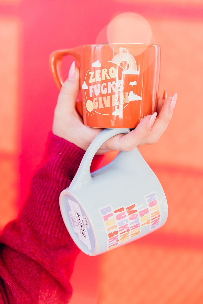 Close of of woman's hand holding two colorful ceramic coffee mugs in her hand in front of a bold pink backdrop. These colorful mugs featuring bold feminist statements can be found at Woman-Owned Wallet, a feminist gift shop in Louisville KY.