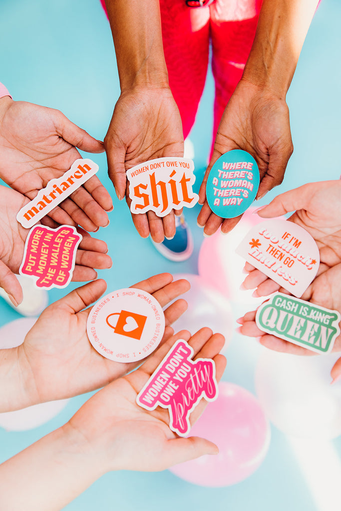 Close up of 4 women's hands holding colorful stickers featuring phrases centering around feminism and money in front of an aqua blue background. These stickers can be found at Woman-Owned Wallet, a feminist gift shop in Louisville KY.