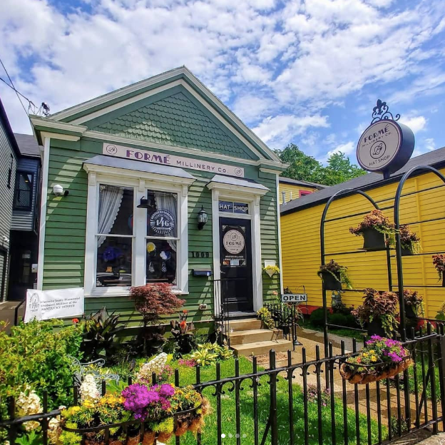 Green shotgun style building with beautiful landscaping, bright green grass, and a cloudy blue sky. This is the Formé Millinery Co. shop, one of the featured businesses on the Nulu Woman-Owned Walking Tour and in the Woman-Owned Directory.
