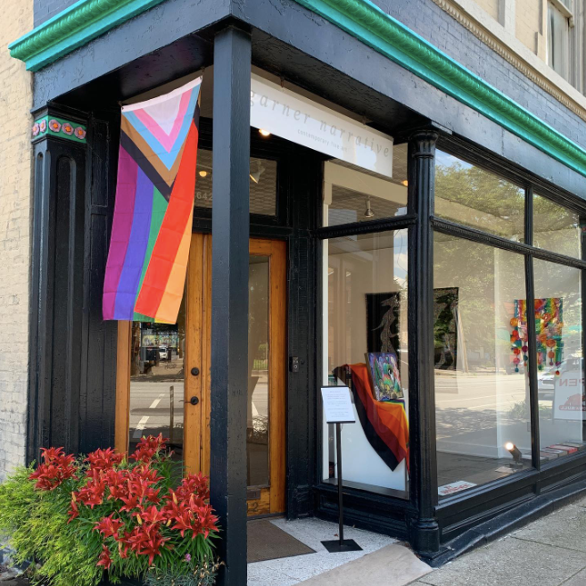 Photo of the Garner Narrative art gallery storefront with wooden double doors, large glass windows showcasing artwork, and an LBGTQIA+ flag hanging in the doorway. This is one of the businesses featured on the Nulu Woman-Owned Walking Tour.
