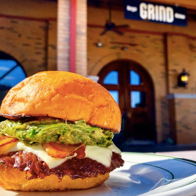 Juicy cheeseburger sitting on a plate with the facade of Grind Burger Kitchen in the background. This is one of the businesses featured on the Nulu Woman-Owned Walking Tour and Woman-Owned Directory hosted by WOW.