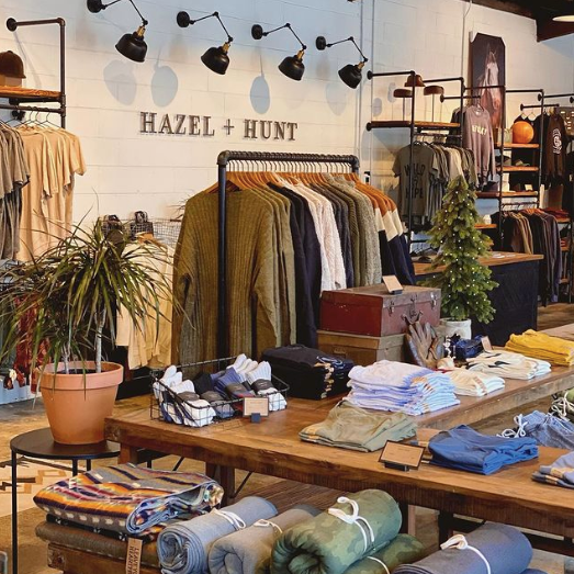 Merch tables and racks full of t-shirts and other apparel at Hazel and Hunt. The back wall is white shiplap with 4 overhead lights shining on the company's logo. This is one of the stops on the Woman-Owned Walking Tour and in the Woman-Owned Directory.