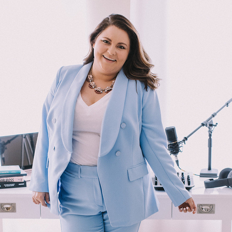 Amanda Dare, founder of Woman-Owned Wallet and host of Woman-Owned Wallet: The Podcast, is posing in front of a desk full of podcast equipment while wearing a light blue power suit and smiling at the camera.