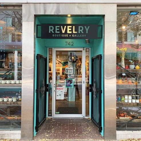 Storefront of Revelry Boutique and Gallery featuring large glass windows and shiny metal trim with a teal blue entryway leading to the front door. There are a variety of products sitting in the front windows. 