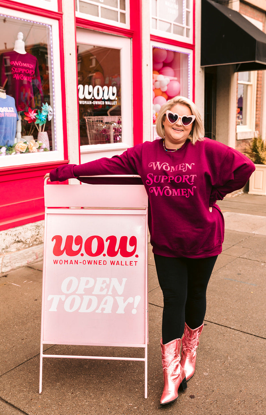 Amanda Dare, owner and founder of Woman-Owned Wallet is standing in front of the bright pink WOW storefront in the Nulu neighborhood of Louisville KY. She is wearing pink heart sunglasses, a WOW signature sweatshirt that says "women support women", and pink shiny cowgirl boots.