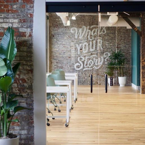 Photo of the Story Louisville coworking space with a brick wall that says "What's Your Story?", white tables with green rolling chairs, wooden floors, and various potted plants. This is one of the businesses on the Nulu Woman-Owned Walking Tour.