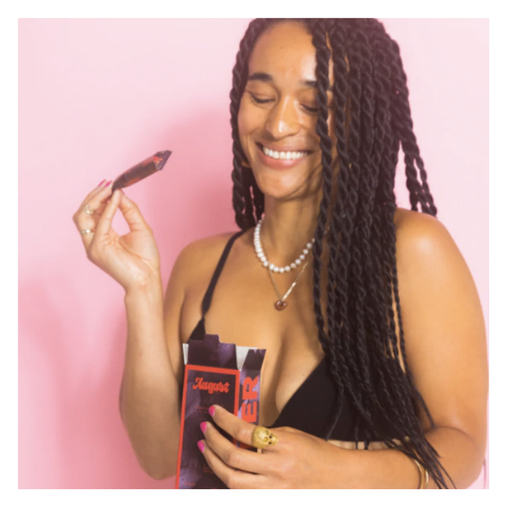 Woman with black braided hair is smiling and holding a box of tampons while standing in from of a pink backdrop. These period care products from August (a brand of color) can be found at Woman-Owned Wallet, a feminist gift shop in Louisville KY.