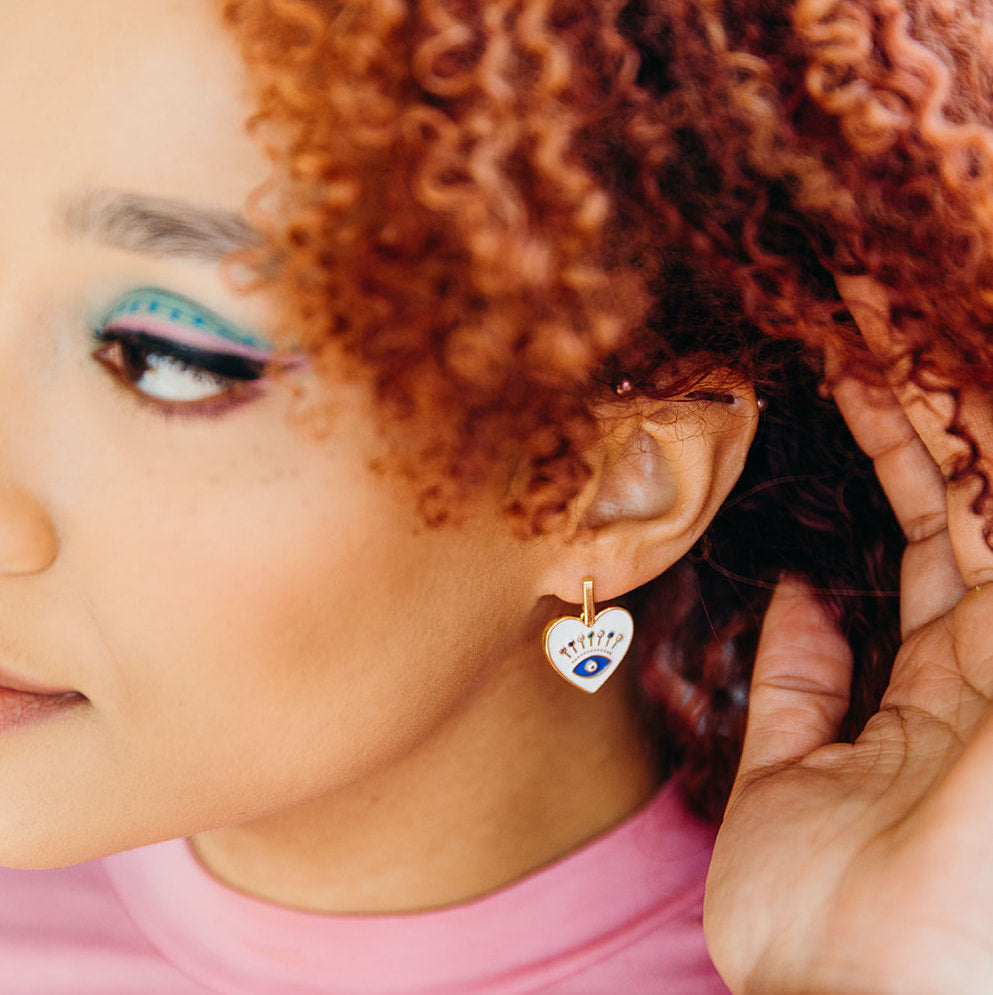 Close-up shot of a woman's face wearing a white heart shaped earring featuring a blue eye in the center. Woman has red curly hair and brightly colored makeup. These earrings, from SassCat Creative, are sold at Woman-Owned Wallet, a feminist gift shop.