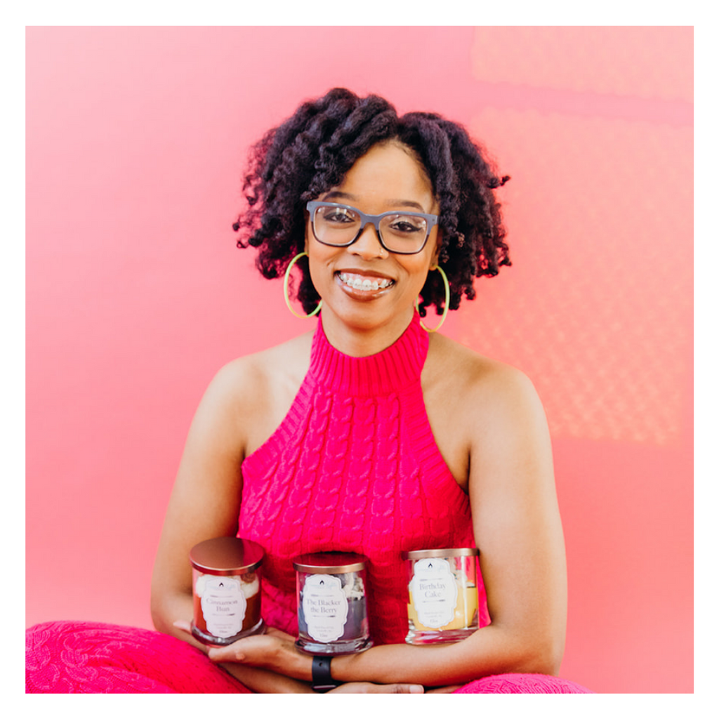 Woman with black coiled hair, glasses and yellow hoop earrings is wearing a hot pink sweater outfit while holding a set of 3 candles. These candles are from a woman-owned brand of color, Serene Nights, and are sold at Woman-Owned Wallet.