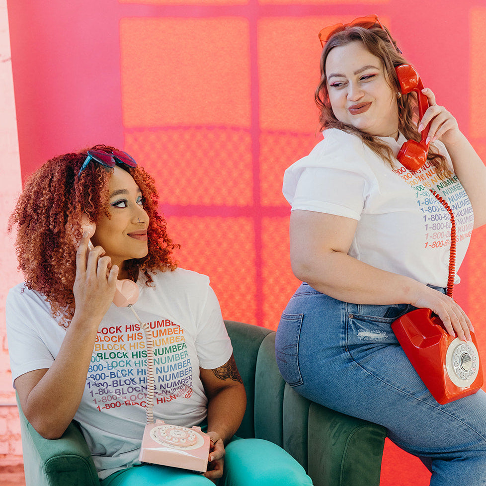 Two women sitting in a vintage chair smiling at each other whole holding rotary phones up to their ears. Both are wearing a white shirt with colorful design that says "1-800-block-his-number". Photoshoot for Woman-Owned Wallet, a feminist gift shop.