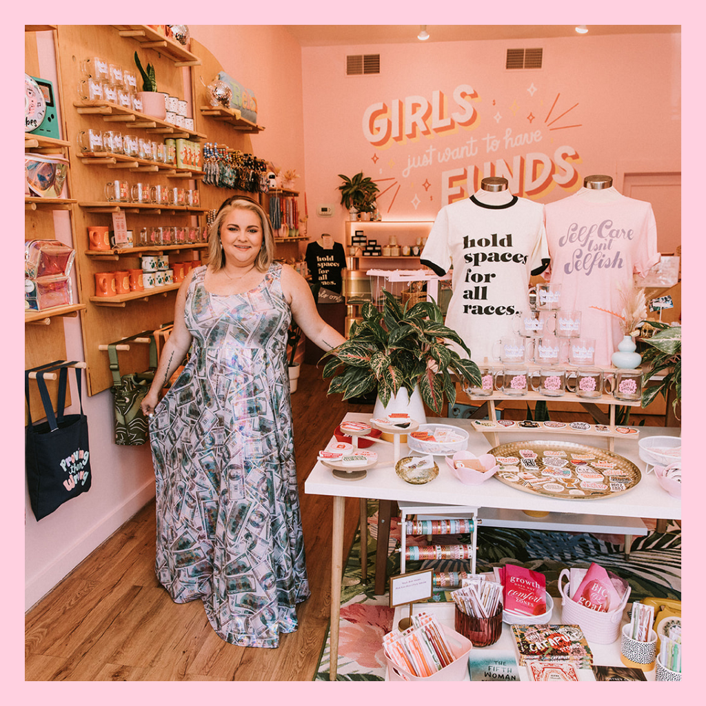 Owned of Woman-Owned Wallet, Amanda Dare, is standing inside the shop posing next to a table full of feminist merchandise. There are wooden shelves behind her that are also full of merchandise. Amanda is wearing a long dress with a dollar bill print.