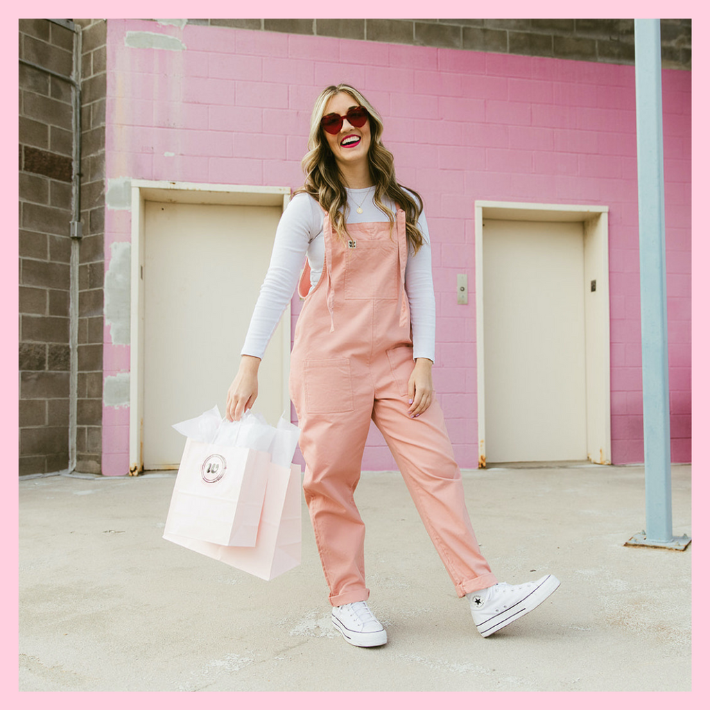 Woman cheerfully looking at the camera wearing pink overalls with a long sleeve white shirt and white sneakers. She is holding two pink gift bags and standing in front of a pink parking garage wall. Photo for Woman-Owned Wallet wholesale page.